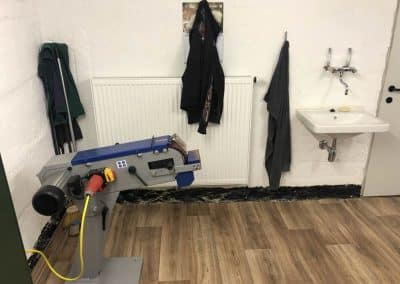 Eine blau, graue Maschine für Waffen, dahinter hängen Jacken an der Wand und gleich daneben befindet sich ein Waschbecken.